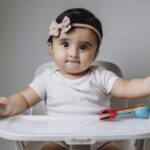 Cute baby girl in high chair with colorful toy utensils against gray background.