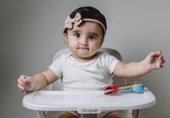 Cute baby girl in high chair with colorful toy utensils against gray background.